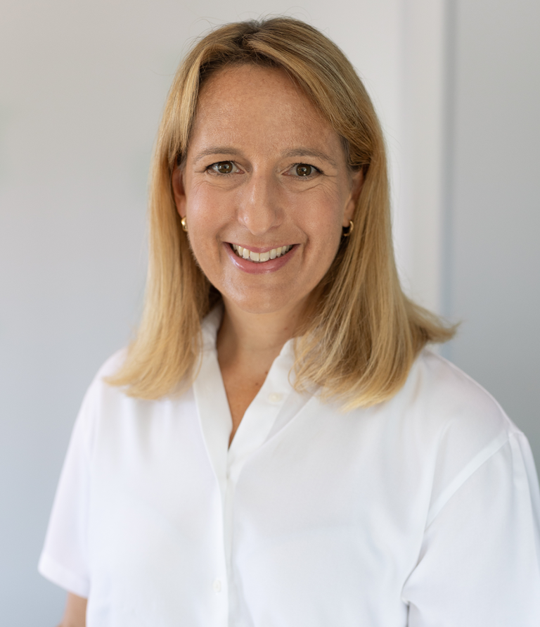 Ein Bild von Dr. med. Elisa Carolus. Lächelnde Frau mit blonden Haaren in einem weißen Hemd vor neutralem Hintergrund.