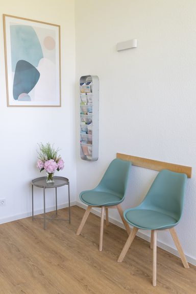 Zwei mintgrüne Stühle stehen an einer hellen Wand, daneben eine Blumenarrangement und Zeitschriftenhalter.
