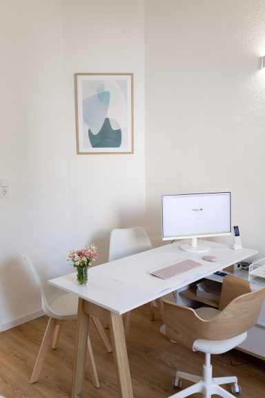 Heller Arbeitsplatz mit Schreibtisch, Computer und einer Blume in der Vase.