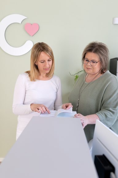 Zwei Frauen besprechen etwas an einem Tisch vor einer hellgrünen Wand.